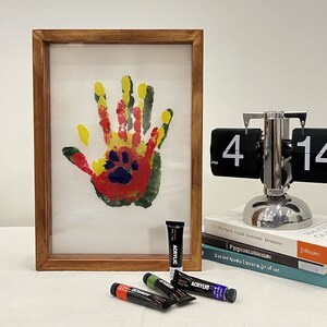 May include: A framed handprint art piece with a paw print in the center, created with red, yellow, green, and blue paint. Several tubes of acrylic paint in various colors are in front of the frame. A silver flip clock displays the time.