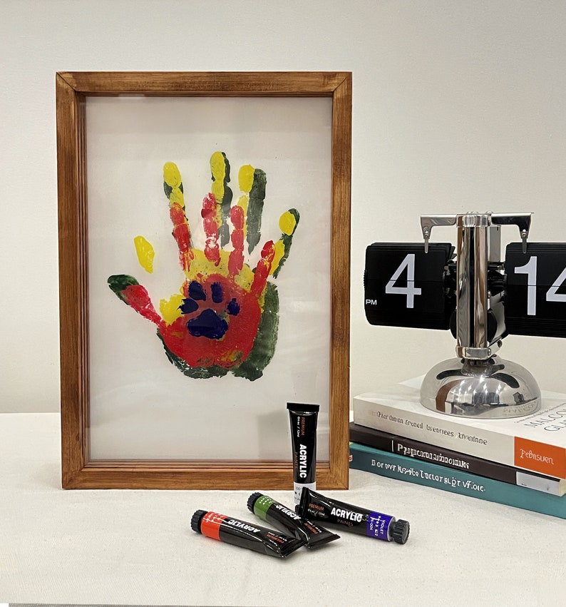 May include: A framed handprint art piece with a paw print in the center, created with red, yellow, green, and blue paint. Several tubes of acrylic paint in various colors are in front of the frame. A silver flip clock displays the time.