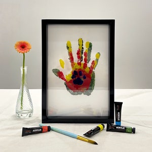 May include: A framed handprint art piece with red, yellow, and green paint, including a paw print. A vase with a single orange flower, paint tubes, and a paintbrush are also present.