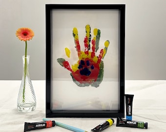 May include: A framed handprint art piece with red, yellow, and green paint, including a paw print. A vase with a single orange flower, paint tubes, and a paintbrush are also present.