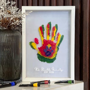 May include: A framed handprint art piece with a white frame. The handprint is a colorful mix of red, yellow, green, and blue, with a paw print in the center. The text "The Smith Family EST. 2025" is printed below the handprint.