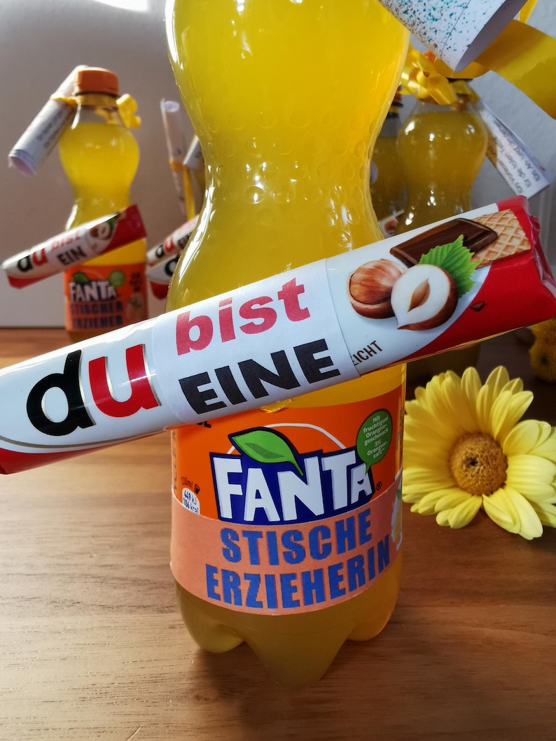 K&ouml;nnte beinhalten: Eine gelbe Flasche Fanta-Limonade mit einem Etikett, auf dem "Du bist eine stiches Erzieherin" (Du bist eine strenge Erzieherin) steht, und eine Schokoladentafel mit einer Haselnuss darauf. Die Schokoladentafel sagt "Du bist eine stich" (Du bist ein Stich).