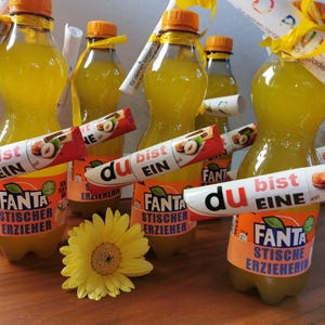 May include: Several bottles of Fanta soda with orange labels and orange caps are arranged on a wooden surface. Each bottle has a candy bar and a rolled-up note attached. A yellow flower is in the foreground.