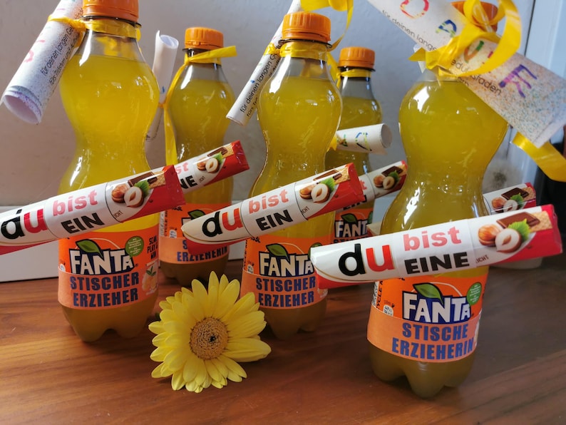 May include: Several bottles of Fanta soda with orange labels and orange caps are arranged on a wooden surface. Each bottle has a candy bar and a rolled-up note attached. A yellow flower is in the foreground.