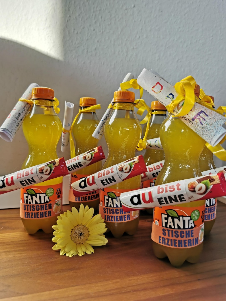 May include: Several bottles of Fanta soda with orange labels and yellow caps are arranged on a wooden surface. Each bottle is adorned with a candy bar and a rolled-up paper tied with a yellow ribbon. A yellow flower is in the foreground.