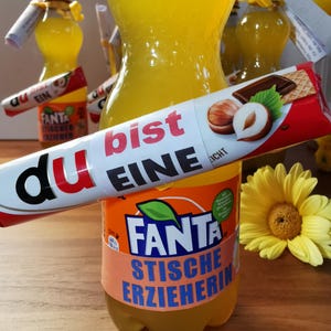 May include: A clear plastic bottle of Fanta soda with a label that reads "STISCHE ERZIEHERIN" and a candy bar with the words "du bist EINE" on it. The image also includes a yellow flower and other bottles of soda.
