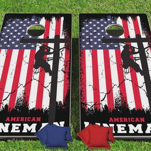 May include: Two rectangular cornhole boards featuring a distressed American flag background with a black silhouette of a utility worker on a pole. The text AMERICAN LINEMAN is printed in white and red at the bottom. Includes blue and red bean bags.