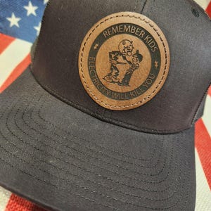 May include: A black baseball cap featuring a circular brown leather patch on the front. The patch displays a graphic of a cartoon character interacting with a child, surrounded by the text REMEMBER KIDS ELECTRICITY WILL KILL YOU.