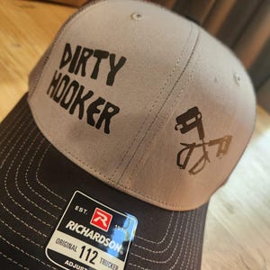 Könnte beinhalten: Eine graue und schwarze Trucker-Cap mit dem Schriftzug DIRTY HOOKER in schwarzen Buchstaben und einer Grafik von zwei gekreuzten Angelhaken. Die Kappe hat einen schwarzen Schirm mit weißen Nähten und einen Aufkleber mit RICHARDSON 112 TRUCKER.