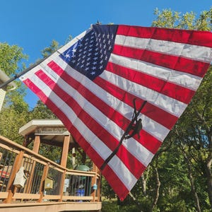 May include: A large American flag featuring thirteen horizontal red and white stripes and a blue canton with white stars. A black silhouette of a utility worker climbing a pole is printed over the stripes. The flag is mounted on a pole outdoors.