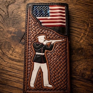 May include: A rectangular leather wallet featuring a hand-tooled basketweave pattern. The front displays a raised, painted figure of a soldier in a dress uniform aiming a rifle, positioned below a painted American flag with red, white, and blue.