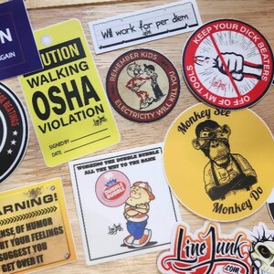 May include: A collection of various vinyl stickers featuring diverse graphic designs, including text-based warnings, cartoon illustrations, and humorous slogans related to electrical line work, all displayed together on a light wooden surface.