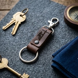 May include: A brown leather keychain featuring a laser-engraved graphic of a utility worker on a power pole. The keychain includes a silver-toned metal lobster clasp and a circular split ring, secured with a single dark metal rivet.