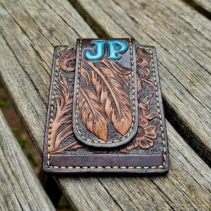 May include: A rectangular brown leather money clip featuring an embossed feather and floral pattern with light-coloured stitching. The top flap displays the letters JP in a bright turquoise blue colour, set against the dark brown textured leather surface.