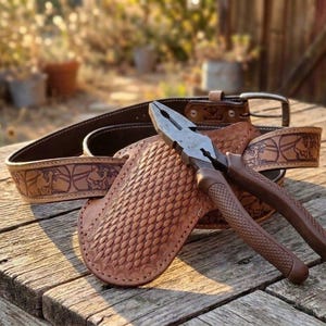 May include: A brown leather belt with a stamped pattern and a matching leather holster featuring a basket-weave texture. A pair of metal pliers with brown textured handles rests on top of the holster, which is positioned in front of the belt.