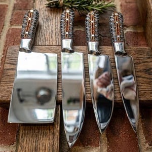 May include: A set of four kitchen knives with polished silver blades and textured, dark-patterned handles featuring metallic rivets. The collection includes a wide cleaver and three varying utility knives, all arranged neatly on a wooden board.