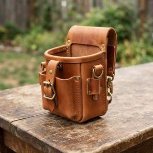 Könnte beinhalten: Eine Werkzeugtasche aus hellbraunem Leder mit strukturierter Öffnung, einer Vordertasche mit Messingschnalle und seitlichen Befestigungspunkten. Die Tasche verfügt über verstärkte Nieten und eine stabile Gürtelschlaufe auf der Rückseite.