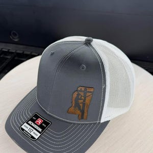 Peut inclure: Une casquette trucker grise et blanche avec un écusson en cuir marron en forme de l'État du Mississippi. L'écusson présente la silhouette d'un ouvrier sur un poteau électrique. Un autocollant noir sur la visière indique Richardson 112 Trucker Adjustable.