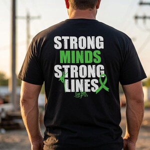 以下が含まれることがあります： 背面に「STRONG MINDS STRONG LINES」という白と緑の文字がプリントされた黒の半袖Tシャツ。デザインには緑色の啓発リボンと送電線作業員の小さなグラフィックが含まれており、その下に「Line Junk」という文字が配置されています。
