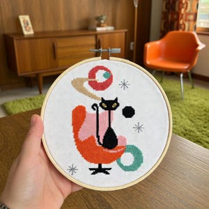 Op de afbeelding: Een kruissteekborduurwerk in een houten hoepel toont een zwarte kat die in een oranje en roze stoel zit. Het ontwerp bevat een rode en bruine planeet, een zwarte stip en turquoise en grijze accenten. De achtergrond is wit.