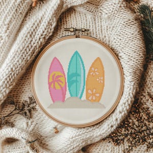 May include: A cross-stitch embroidery hoop featuring three colorful surfboards. The surfboards are pink with a palm tree, teal with a leaf design, and orange with flowers. The design is set against a sand-colored background.