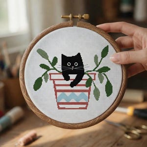 Op de afbeelding: Een kruissteek borduurring met een zwarte kat in een rood en blauw gestreepte pot met groene bladeren. De borduursel zit in een houten ring met een messing sluiting.