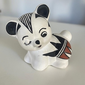 Puede incluir: Figura de oso de cerámica en una pose juguetona, pintada en blanco con patrones geométricos negros y rojos. El oso tiene un ojo cerrado en un guiño, orejas negras y un diseño a rayas blanco y negro en la cabeza.