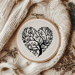 May include: A black and white cross-stitch design of a tree in the shape of a heart, set in a wooden embroidery hoop. The artwork is displayed on a cream-colored, knitted fabric background.