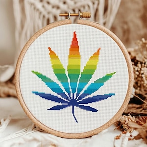 May include: A cross-stitch embroidery hoop featuring a colorful cannabis leaf design. The leaf is rendered in a gradient of rainbow colors, from red to blue, against a white background. The wooden hoop is visible.