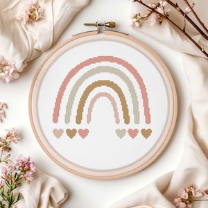 May include: A cross-stitch rainbow design in a wooden embroidery hoop. The rainbow features pastel colors: pink, beige, and gold. Small heart shapes are at the bottom of the rainbow. The background is white.