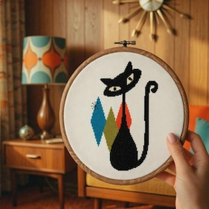 May include: A cross-stitch embroidery of a black cat with a long tail, set against a white background. The cat has a stylized design with blue, green, and orange diamond shapes. The embroidery is framed in a round wooden hoop.