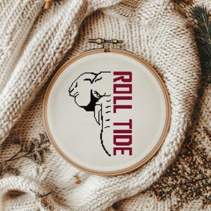 May include: A cross-stitch embroidery hoop featuring a black and white elephant design with the words "ROLL TIDE" in maroon. The hoop is set against a cream-colored fabric, with a cozy, textured background.