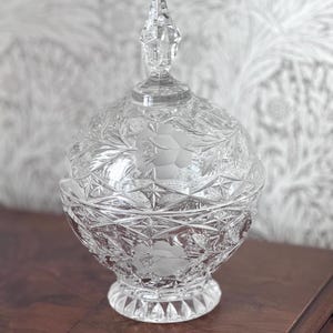 May include: A clear, cut-glass candy dish with a lid. The round dish features etched floral designs and geometric patterns. The lid is topped with a pointed finial. The dish sits on a pedestal base.
