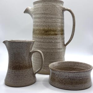 May include: A ceramic coffee set comprising a tall, lidded coffee pot, a creamer jug, and a small bowl. The set has a speckled, off-white and brown glaze. The coffee pot has a curved handle and spout. The creamer has a handle and a flared top. The bowl is round.