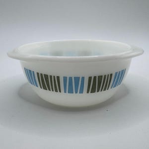 May include: White glass bowl with a rounded shape and a wide rim. The bowl features a pattern of vertical blue and green stripes around the exterior. The bowl is likely vintage and suitable for serving or display.