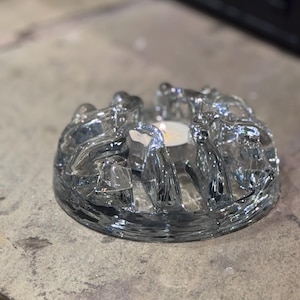 May include: A clear glass candle holder with a circular design. The holder has curved, raised sections around the edge, with openings for tea lights. A lit tea light is visible inside, casting a warm glow.