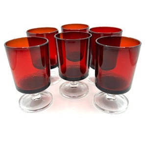 May include: A set of six vintage red wine glasses. Each glass has a ruby red bowl and a clear, circular base. The glasses are arranged in a semi-circle, showcasing their elegant design. Ideal for serving wine at a dinner party or special occasion.