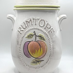 May include: A white ceramic Rumtopf jar with a ribbed texture, two side handles, and a matching lid. The front features a raised, oval-shaped relief of a peach and a plum with green leaves, surrounded by the embossed text RUMTOPF in capital letters.