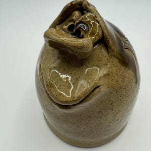 May include: A handmade ceramic jar with a quirky face design. The jar is a mottled brown colour with darker brown accents, featuring a smiling face with closed eyes and a hand covering the top. The jar has a rounded body and a flat base.