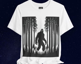 May include: White t-shirt with a black silhouette of Bigfoot standing in a forest. The design is centered on the shirt.