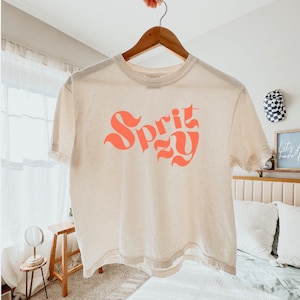 May include: A cream coloured cropped t-shirt with a pink graphic that says "Spritzzy".