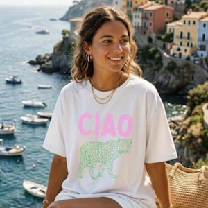 May include: White t-shirt with the word "CIAO" in pink block letters and a green leopard design. The shirt is worn by a person with layered gold necklaces and hoop earrings. The background features a coastal town with colorful buildings and boats.