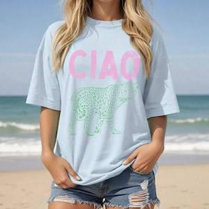 May include: Light blue t-shirt with the word "CIAO" in pink block letters above a green leopard print design. The shirt has short sleeves and a relaxed fit. The background shows a beach and ocean.