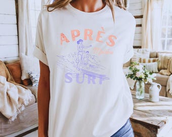 Könnte beinhalten: Ein cremefarbenes Kurzarm-T-Shirt mit einem Grafikdruck und dem Text APRÈS SURF Malibu. Das Design zeigt eine Strichzeichnung von zwei Personen, die auf einer Welle surfen, wobei das Wort APRÈS in Orange und SURF in Hellviolett gehalten ist.