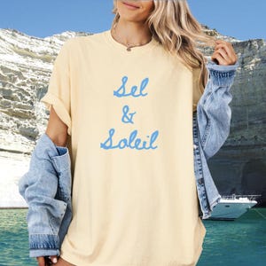 Puede incluir: Una camiseta amarilla clara con las palabras "Sel & Soleil" en escritura azul. La camiseta se usa con una chaqueta vaquera. El fondo presenta un paisaje marino con un barco.