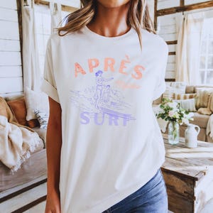 May include: Cream-colored t-shirt with the words "APRÈS Malibu SURF" in coral and purple. The graphic features a surfer on a wave. The shirt has short sleeves and a relaxed fit, perfect for casual wear.