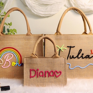 Puede incluir: Tres bolsas de mano de yute con asas marrones. La bolsa más grande tiene un arcoíris y la letra "B". La bolsa mediana tiene el nombre "Tulia" con una palmera y un sol. La bolsa más pequeña tiene el nombre "Diana" con un corazón.
