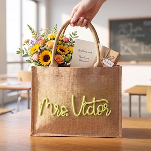 May include: A tan burlap tote bag with two handles featuring the name Mrs. Victor in yellow cursive lettering. Inside the bag is a bouquet of sunflowers and roses, a white card reading Gratitude card Thank you, Professor!, and a gift box with a tag.