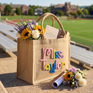 May include: A tan burlap tote bag featuring the text "miss. Taylor" in raised, multi-colored yarn lettering. The bag contains a bouquet of sunflowers, white roses, and purple lavender, along with a card that reads "thank you GRATITUDE".