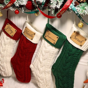 May include: Four personalised Christmas stockings in red, white, and green, with names on leather tags. The stockings are knitted with a cable pattern and have white cuffs. Festive holiday decor with ribbons and ornaments is visible in the background.